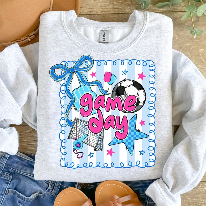 May include: A light gray sweatshirt with a colorful "Game Day" graphic. The design includes a soccer ball, stars, a bow, and the words "Game Day" in pink and blue. The sweatshirt is a Gildan brand.