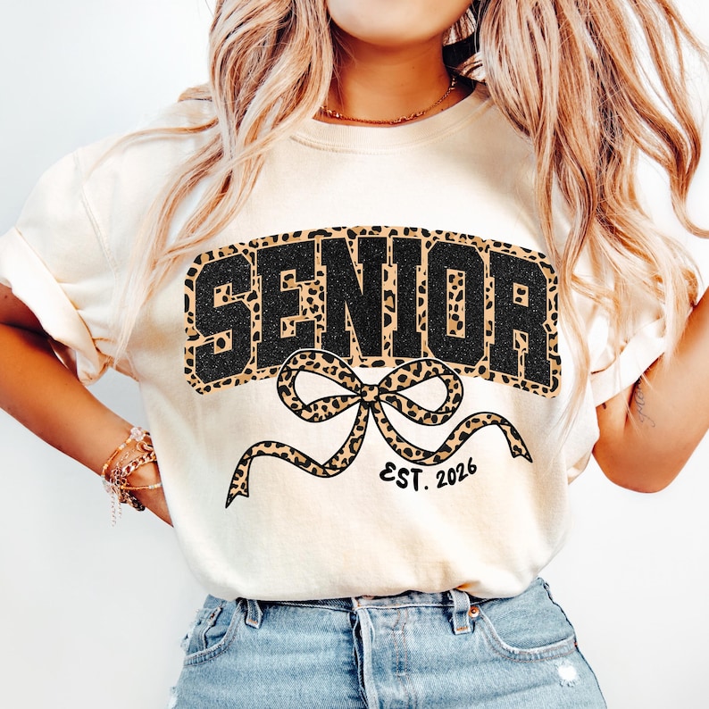 May include: Cream-colored t-shirt with the word "SENIOR" in black block letters with a leopard print fill. A leopard print bow is below the word, with "EST. 2026" in a smaller font.