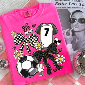 May include: A hot pink t-shirt featuring a soccer-themed design. The graphic includes a soccer jersey with the number 7, soccer balls, bows, daisies, and a soccer glove. The shirt is a Comfort Colors brand, size L-G.