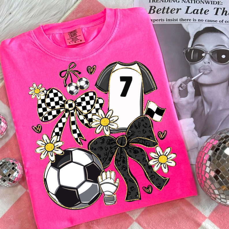 May include: A hot pink t-shirt featuring a soccer-themed design. The graphic includes a soccer jersey with the number 7, soccer balls, bows, daisies, and a soccer glove. The shirt is a Comfort Colors brand, size L-G.