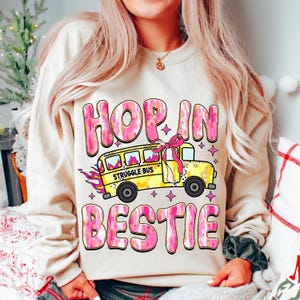 May include: A cream-colored sweatshirt with a pink and gold graphic design. The graphic features a yellow school bus with the words "Struggle Bus" on the side, a pink bow, and the text "Hop in Bestie".