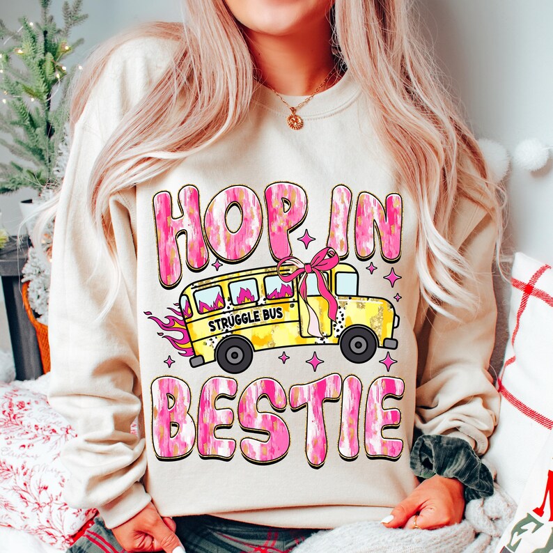 May include: A cream-colored sweatshirt with a pink and gold graphic design. The graphic features a yellow school bus with the words "Struggle Bus" on the side, a pink bow, and the text "Hop in Bestie".