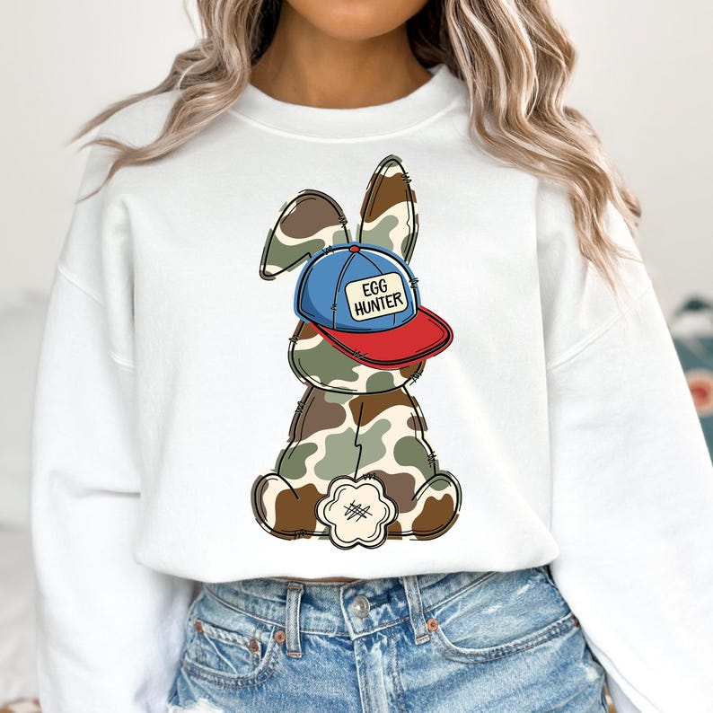 May include: White sweatshirt with a cartoon bunny wearing a blue and red baseball cap that says "Egg Hunter". The bunny is wearing camouflage.
