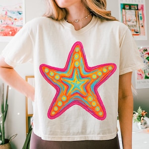May include: Cream-colored t-shirt featuring a vibrant, multi-colored starfish design. The starfish is outlined in hot pink, with orange, yellow, and blue hues filling the interior. Small yellow circles are evenly spaced around the starfish's edges.