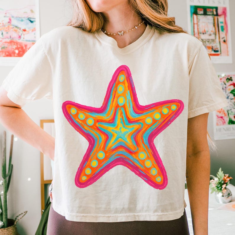 May include: Cream-colored t-shirt featuring a vibrant, multi-colored starfish design. The starfish is outlined in hot pink, with orange, yellow, and blue hues filling the interior. Small yellow circles are evenly spaced around the starfish's edges.