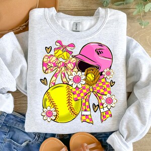 May include: A light gray sweatshirt featuring a softball-themed design. The design includes a pink softball helmet, a yellow softball, a brown glove, and decorative bows and flowers in pink and yellow. The design is embellished with hearts.
