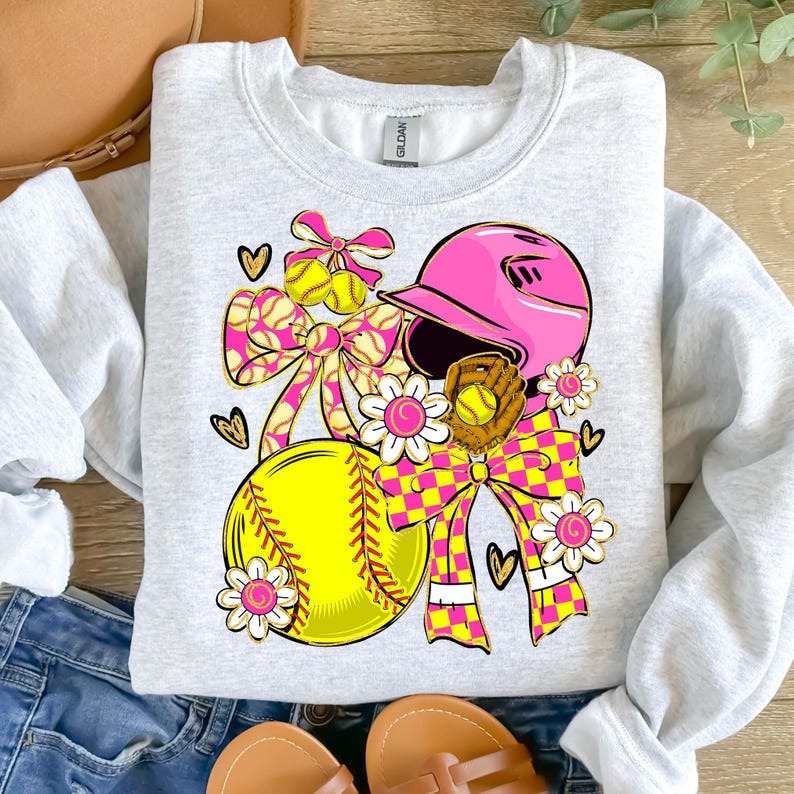 May include: A light gray sweatshirt featuring a softball-themed design. The design includes a pink softball helmet, a yellow softball, a brown glove, and decorative bows and flowers in pink and yellow. The design is embellished with hearts.