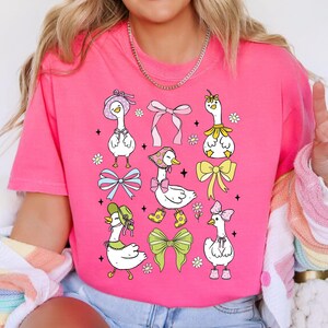 Puede incluir: Una camiseta rosa con un dise&ntilde;o gr&aacute;fico de varios gansos de dibujos animados que llevan sombreros y lazos. Los gansos est&aacute;n rodeados de flores y lazos.