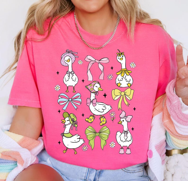 Puede incluir: Una camiseta rosa con un dise&ntilde;o gr&aacute;fico de varios gansos de dibujos animados que llevan sombreros y lazos. Los gansos est&aacute;n rodeados de flores y lazos.