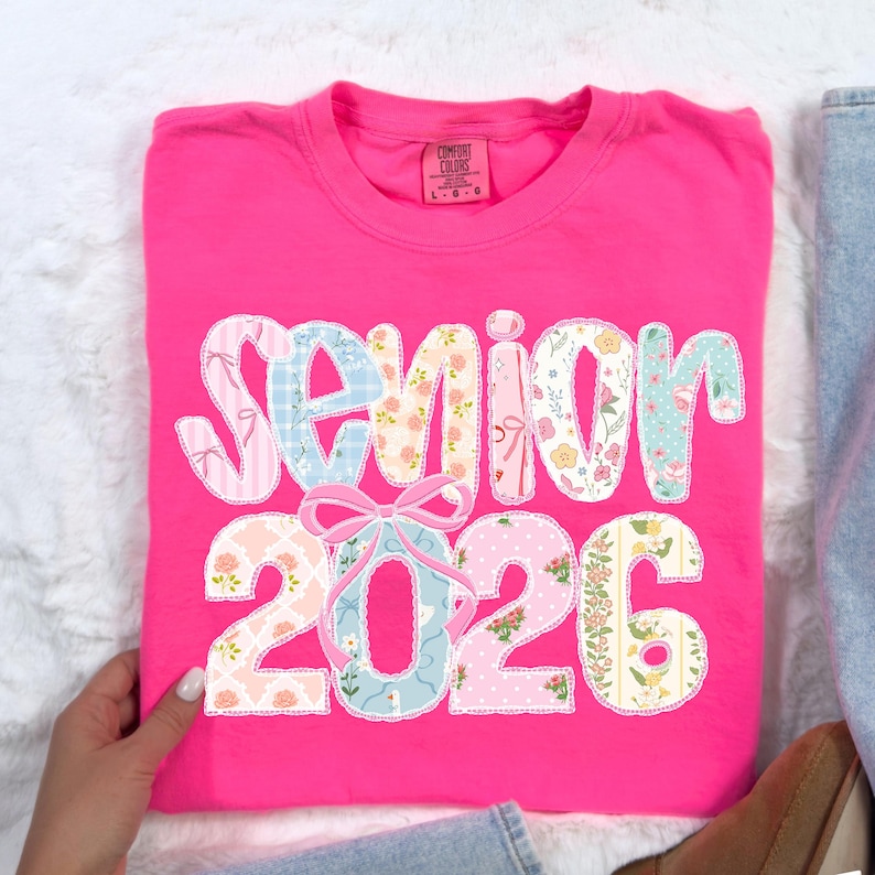 Puede incluir: Una camiseta rosa intenso con la palabra "Senior 2026" en un dise&ntilde;o de patchwork. Las letras y los n&uacute;meros est&aacute;n decorados con telas florales y estampadas, incluyendo lazos. La camiseta est&aacute; doblada sobre una superficie blanca.