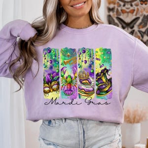 May include: A lavender sweatshirt with a colorful Mardi Gras design featuring a mask, a lobster, a king cake, and a hat. The text "Mardi Gras" is printed below the design.