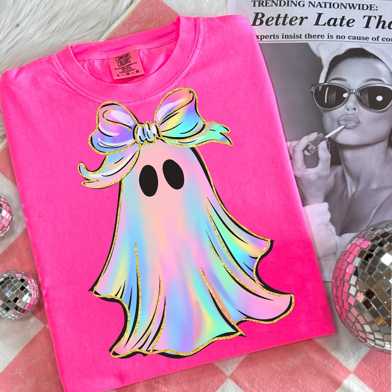 May include: A bright pink sweatshirt featuring a ghost design with a colorful bow and gold glitter accents. The ghost has a shimmering, iridescent appearance. The shirt has the Comfort Colors brand tag. A newspaper clipping is in the background.