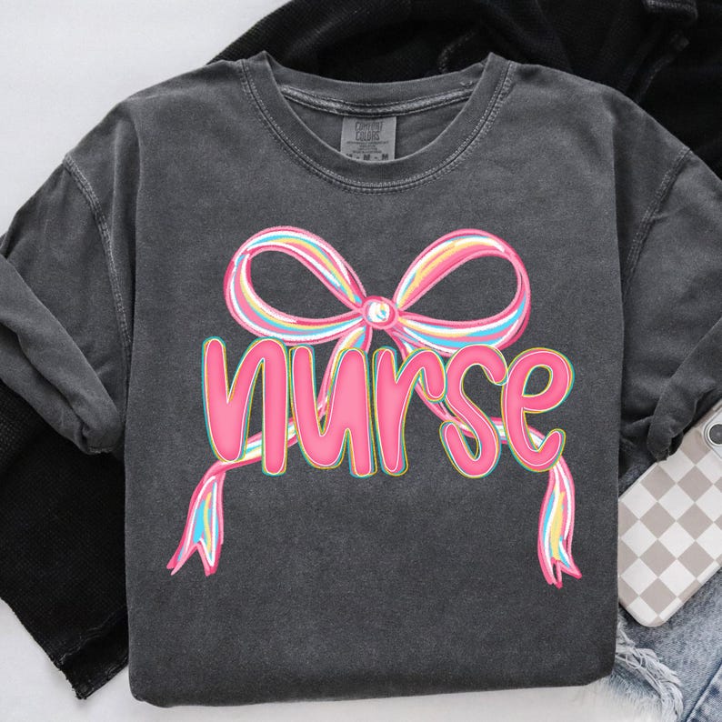 Pode incluir: Camisola cinza escuro com a palavra "Nurse" em rosa, com um la&ccedil;o colorido por cima. O la&ccedil;o tem listras rosa, amarelas, azuis e verdes. A camisola tem mangas compridas e gola redonda.