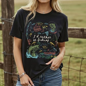 May include: Black t-shirt with a colorful fishing-themed graphic. The design includes illustrations of fish, fishing rods, a tackle box, and the phrase "I'd rather be fishing."