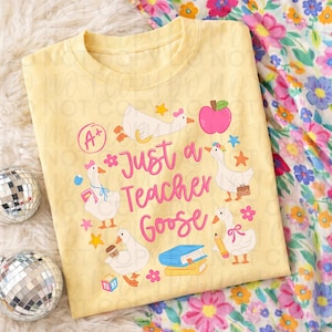 以下が含まれることがあります： ピンク色の「Just a Teacher Goose」という文字が入った、薄い黄色のTシャツ。学校をテーマにしたアクセサリーを身に着けた漫画のガチョウ、ピンクのリンゴ、星、花がデザインされています。シャツは花柄の生地の上に折りたたまれています。