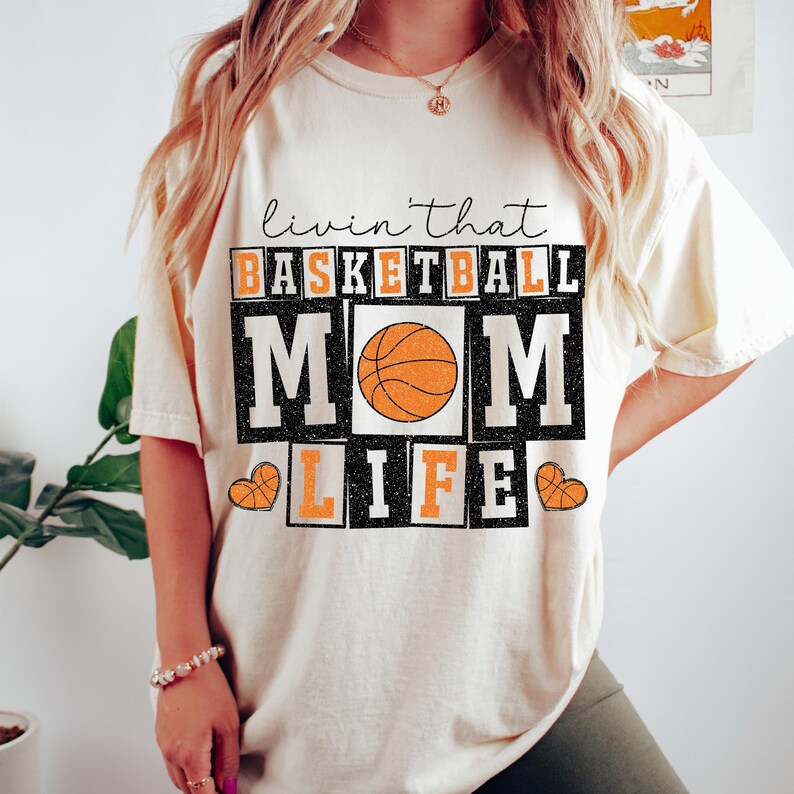 May include: A cream-colored t-shirt with the text "Livin' that Basketball Mom Life" in black and orange lettering. A basketball graphic is in the center of the text.