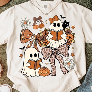 Op de afbeelding: Cr&egrave;mekleurig T-shirt met een Halloween-thema. Het ontwerp bevat twee stripfiguurgeesten die boeken lezen, omringd door strikken, bloemen, pompoenen, sterren en een zwarte kat. De tekst "Comfort Colors" staat bovenaan.