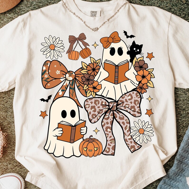 Op de afbeelding: Cr&egrave;mekleurig T-shirt met een Halloween-thema. Het ontwerp bevat twee stripfiguurgeesten die boeken lezen, omringd door strikken, bloemen, pompoenen, sterren en een zwarte kat. De tekst "Comfort Colors" staat bovenaan.