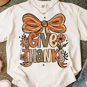 May include: Cream-colored t-shirt featuring the words "GIVE THANKS" in a fall-themed design. The design includes a large bow, flowers, and leaves in shades of orange, brown, and green. A necklace is visible at the neckline.