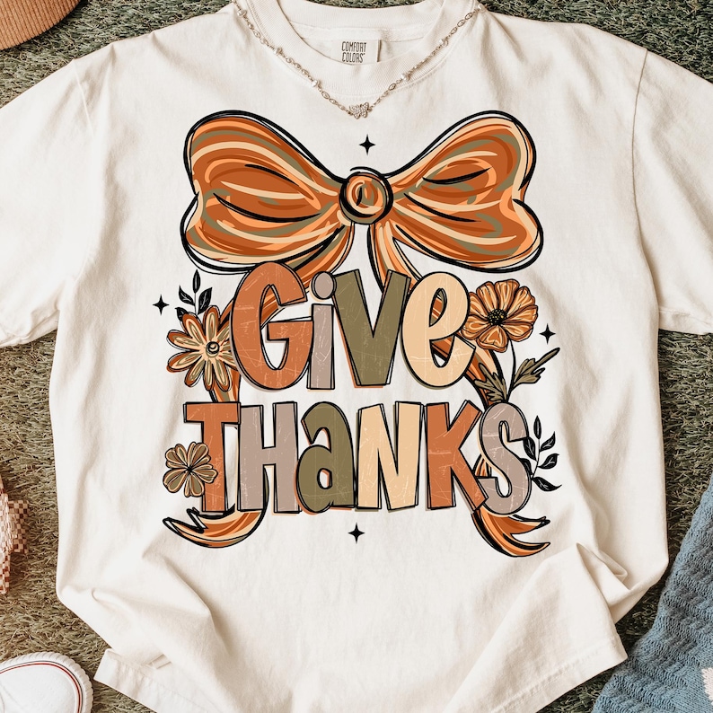 May include: Cream-colored t-shirt featuring the words "GIVE THANKS" in a fall-themed design. The design includes a large bow, flowers, and leaves in shades of orange, brown, and green. A necklace is visible at the neckline.
