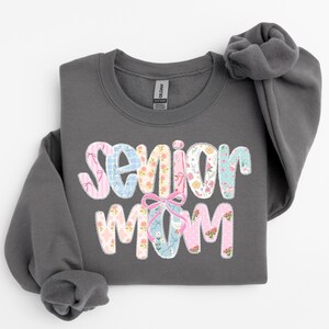 Puede incluir: Una sudadera gris oscuro con las palabras "Senior Mom" en un dise&ntilde;o floral y estampado colorido. El texto presenta patrones rosas, azules y blancos con un lazo rosa. La sudadera tiene cuello redondo y mangas largas.