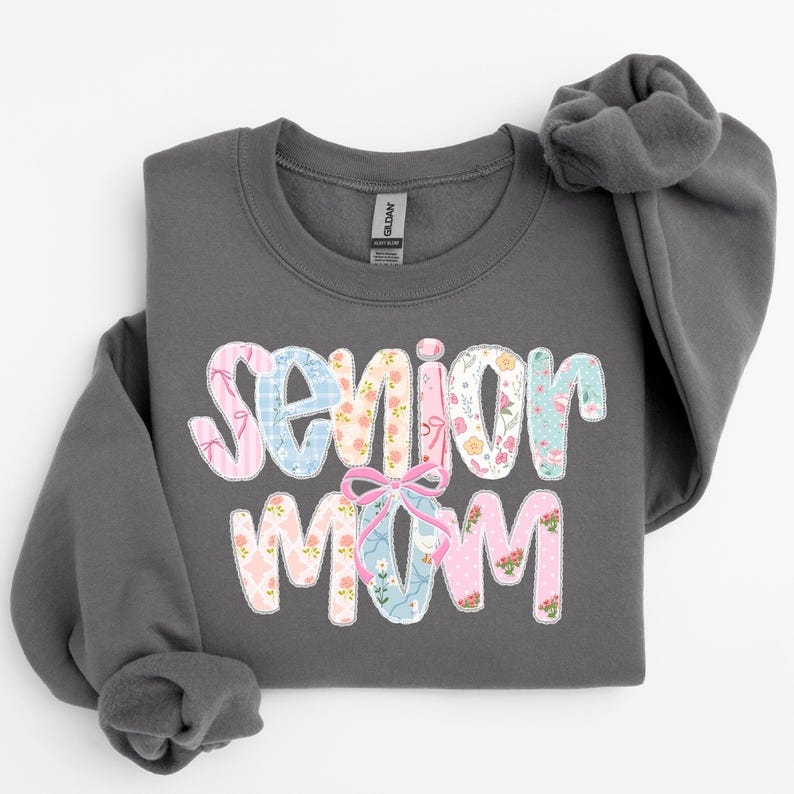 Puede incluir: Una sudadera gris oscuro con las palabras "Senior Mom" en un dise&ntilde;o floral y estampado colorido. El texto presenta patrones rosas, azules y blancos con un lazo rosa. La sudadera tiene cuello redondo y mangas largas.