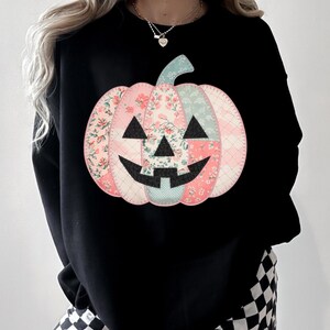 May include: Black sweatshirt featuring a patchwork pumpkin design. The pumpkin is composed of pink and blue floral fabric pieces, with a black face. The stem is light blue. A festive fall-themed apparel item.
