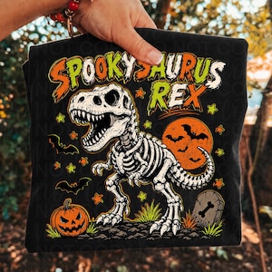 May include: A black t-shirt featuring a graphic of a white T-Rex skeleton. The text reads SPOOKYSAURUS REX in orange and green. The design includes a bright orange full moon with black bats, a carved jack-o'-lantern, a tombstone, and small stars.