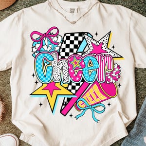 Op de afbeelding: Cr&egrave;mekleurig T-shirt met een levendig grafisch ontwerp met het woord "Cheer" in vetgedrukte, kleurrijke letters. Het ontwerp bevat sterren, een megafoon en een strik, allemaal in helder roze, blauw en geel.