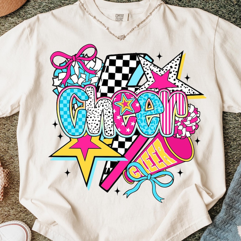 Op de afbeelding: Cr&egrave;mekleurig T-shirt met een levendig grafisch ontwerp met het woord "Cheer" in vetgedrukte, kleurrijke letters. Het ontwerp bevat sterren, een megafoon en een strik, allemaal in helder roze, blauw en geel.