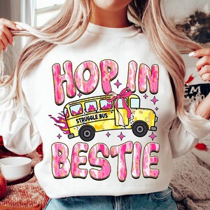 May include: White sweatshirt with a pink and yellow graphic design. The graphic features a yellow school bus with the words "Struggle Bus" on the side. The text "Hop in bestie" is written in pink and yellow letters.
