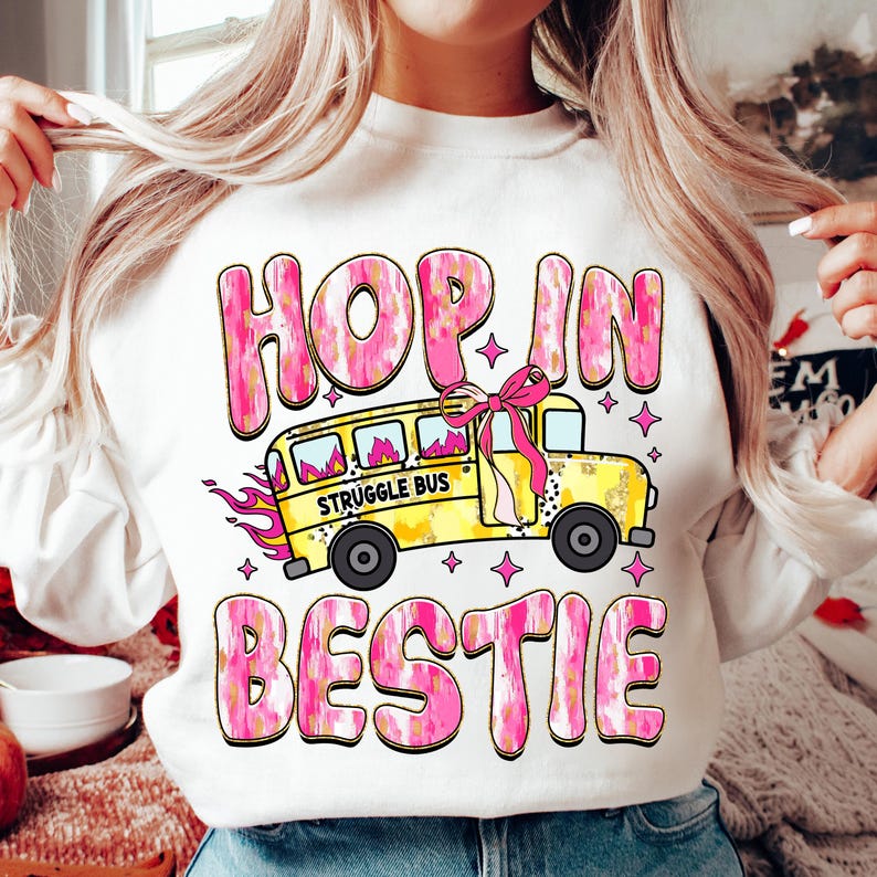 May include: White sweatshirt with a pink and yellow graphic design. The graphic features a yellow school bus with the words "Struggle Bus" on the side. The text "Hop in bestie" is written in pink and yellow letters.
