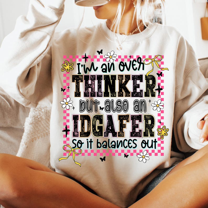 May include: A beige sweatshirt with the text "I'm an over THINKER but also an IDGAFER so it balances out." The text is in black, pink, and green, with floral and butterfly accents.