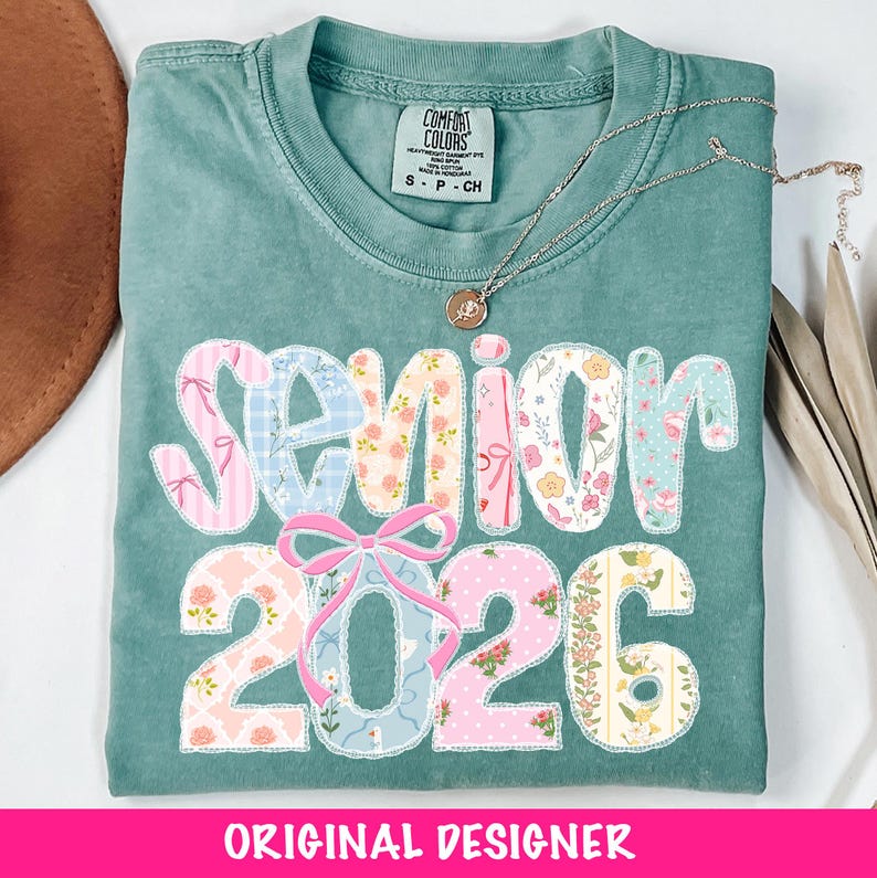 Puede incluir: Una camiseta Comfort Colors verde azulado con la palabra "SENIOR 2026" en un dise&ntilde;o de patchwork. Las letras y los n&uacute;meros est&aacute;n decorados con patrones florales y lazos. Un collar plateado con un colgante descansa sobre la camisa.
