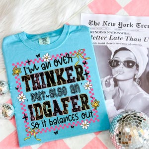 May include: A light blue t-shirt with the text "I'm an over THINKER but also an IDGAFER so it balances out." The graphic includes flowers, butterflies, and bows. The shirt is on a pink and white checkered surface.
