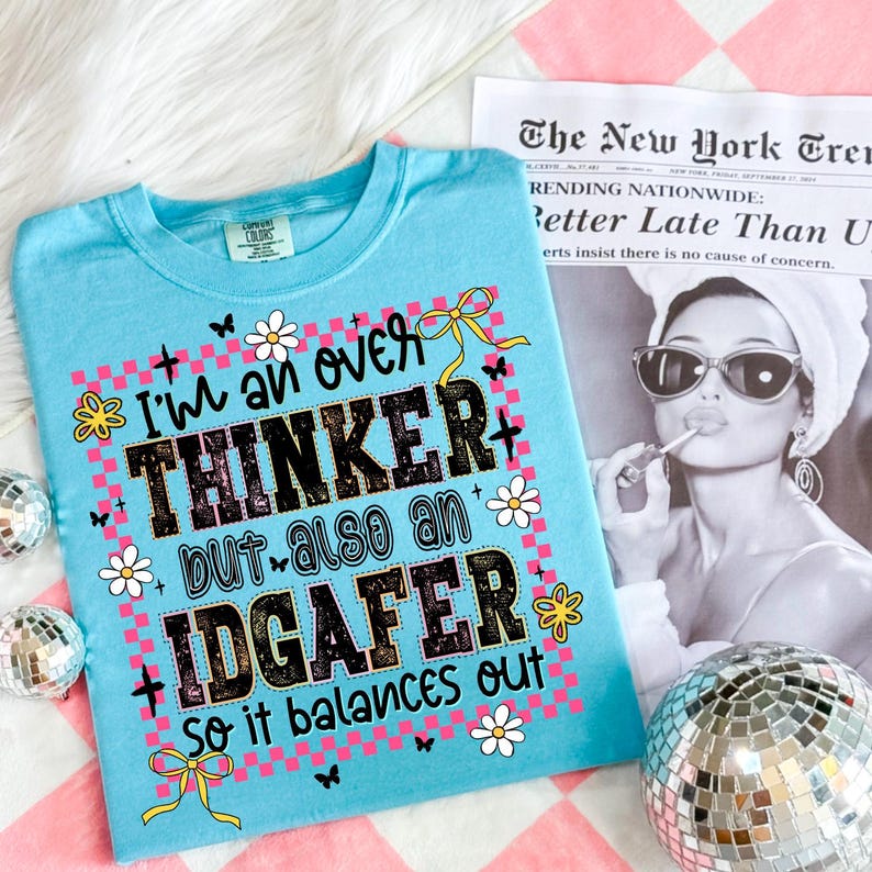 May include: A light blue t-shirt with the text "I'm an over THINKER but also an IDGAFER so it balances out." The graphic includes flowers, butterflies, and bows. The shirt is on a pink and white checkered surface.