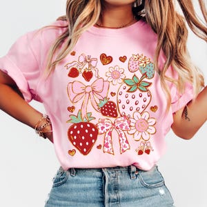 Op de afbeelding: Roze T-shirt met een grafisch ontwerp van aardbeien, strikken en harten. Het ontwerp is in goud, rood, roze en groen.