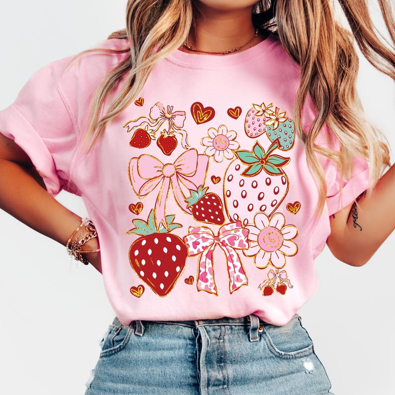 Op de afbeelding: Roze T-shirt met een grafisch ontwerp van aardbeien, strikken en harten. Het ontwerp is in goud, rood, roze en groen.