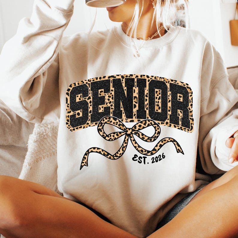 May include: Cream-colored sweatshirt with the word "SENIOR" in black block letters with a leopard print background. A leopard print bow with the text "EST. 2026" is below the word "SENIOR".