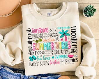 May include: Cream-colored sweatshirt with the words "Summer Vibes" in a colorful, block-letter design. The design includes summer-themed words like "sunshine," "flip flops," and "swimming." The sweatshirt is paired with blue jeans and a tan hat.