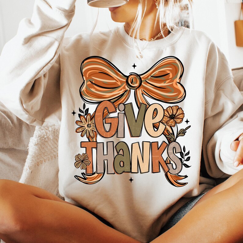 May include: Cream-colored sweatshirt with a large graphic that says "Give Thanks" in a fall-themed color palette. The design includes a bow, flowers, and leaves. The sweatshirt is a long-sleeved, casual top.
