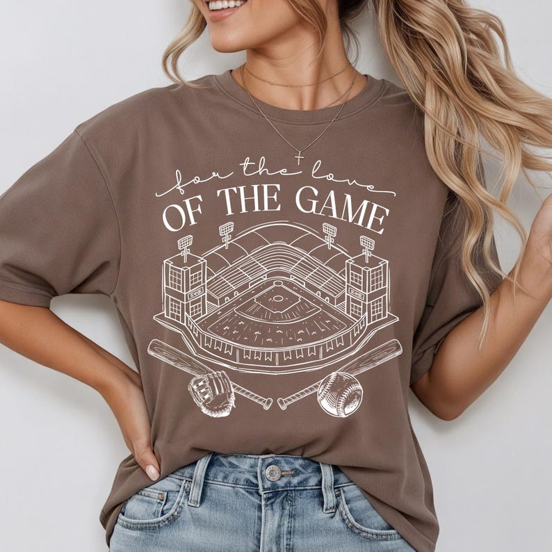 Op de afbeelding: Bruin t-shirt met een witte honkbalstadionafbeelding, honkbal, handschoen en knuppels. De tekst "for the love of the game" staat boven het stadionontwerp. Het shirt is gemaakt van een zacht materiaal.