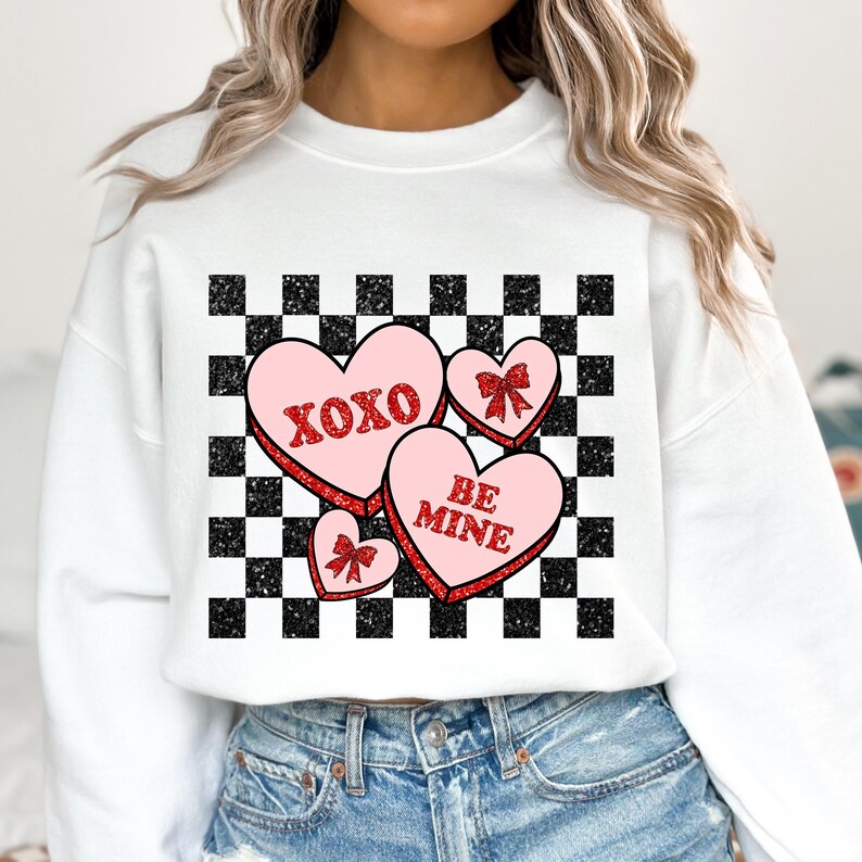 May include: White sweatshirt with a checkered pattern and three pink candy hearts with red glitter. The hearts say "XOXO", "BE MINE", and have a red bow.