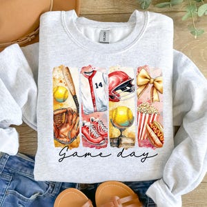May include: A gray sweatshirt with a graphic design of a baseball game. The design includes a baseball bat, glove, ball, jersey, helmet, popcorn, hot dog, and a bow. The text "Game day" is printed below the graphic.