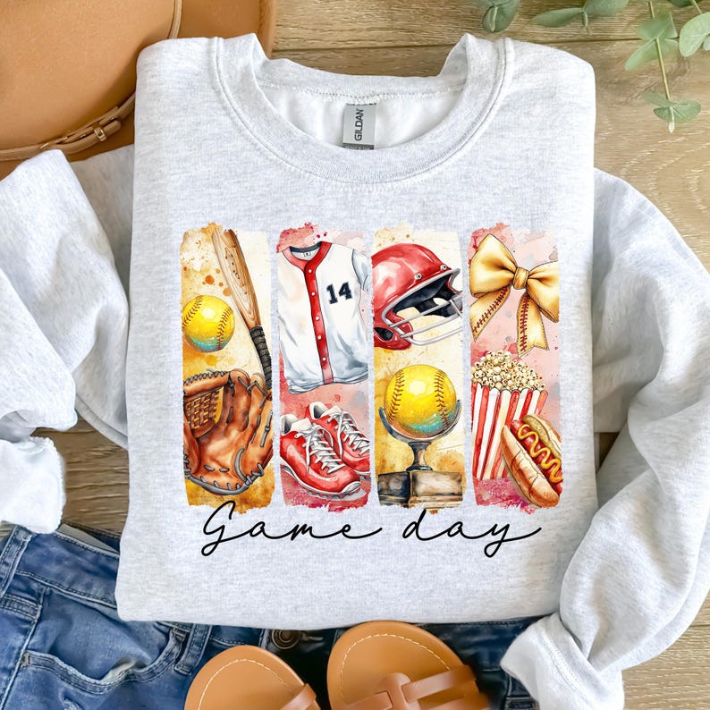May include: A gray sweatshirt with a graphic design of a baseball game. The design includes a baseball bat, glove, ball, jersey, helmet, popcorn, hot dog, and a bow. The text "Game day" is printed below the graphic.