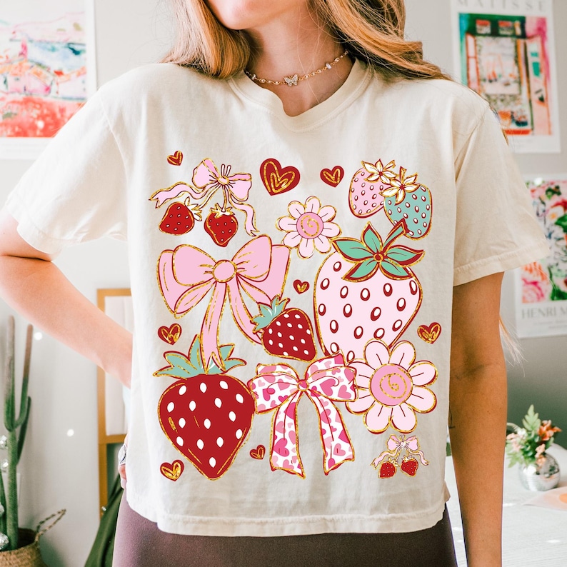 Op de afbeelding: Een wit T-shirt met een grafische print van aardbeien, strikken, harten en bloemen in de kleuren roze, rood, groen en goud.