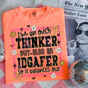 May include: A neon orange t-shirt with the text "I'm an over THINKER but also an IDGAFER so it balances out." The design includes daisies, butterflies, and yellow bows. The shirt is a Comfort Colors brand.