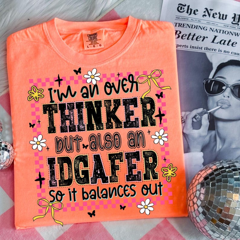 May include: A neon orange t-shirt with the text "I'm an over THINKER but also an IDGAFER so it balances out." The design includes daisies, butterflies, and yellow bows. The shirt is a Comfort Colors brand.