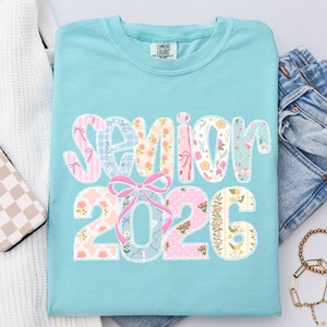 Puede incluir: Una camiseta azul claro con la palabra "SENIOR 2026" en un dise&ntilde;o de patchwork. Las letras y los n&uacute;meros est&aacute;n rellenos de telas florales y estampadas, y un lazo rosa aparece en el dise&ntilde;o.