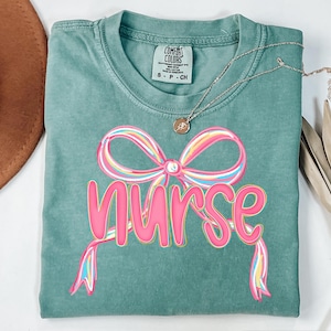 Pode incluir: Uma camiseta Comfort Colors verde-azulado com um design de la&ccedil;o rosa e arco-&iacute;ris e a palavra "nurse" em rosa. Um colar de ouro com um pequeno pingente est&aacute; drapeado sobre a camisa.